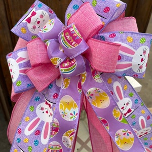 May include: A pink and purple Easter bow with a bunny and egg design. The bow is made of two different ribbons, one with a pink and white polka dot pattern and the other with a purple and white polka dot pattern. The bow is tied with a pink ribbon and has a small bow on top. The bow is decorated with a bunny and egg design. The bow is perfect for adding a touch of Easter cheer to your home.