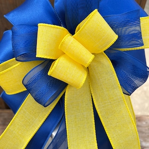 May include: A blue and yellow bow with a wired edge. The bow is made of two different types of ribbon, one is a solid color and the other is a sheer ribbon with a striped pattern. The bow is tied with a knot in the center.