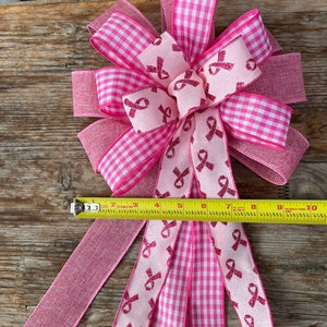 May include: Pink and white gingham ribbon bow with pink ribbon accents featuring a pink ribbon design. The bow is tied with a pink burlap ribbon and measures approximately 10 inches in length.
