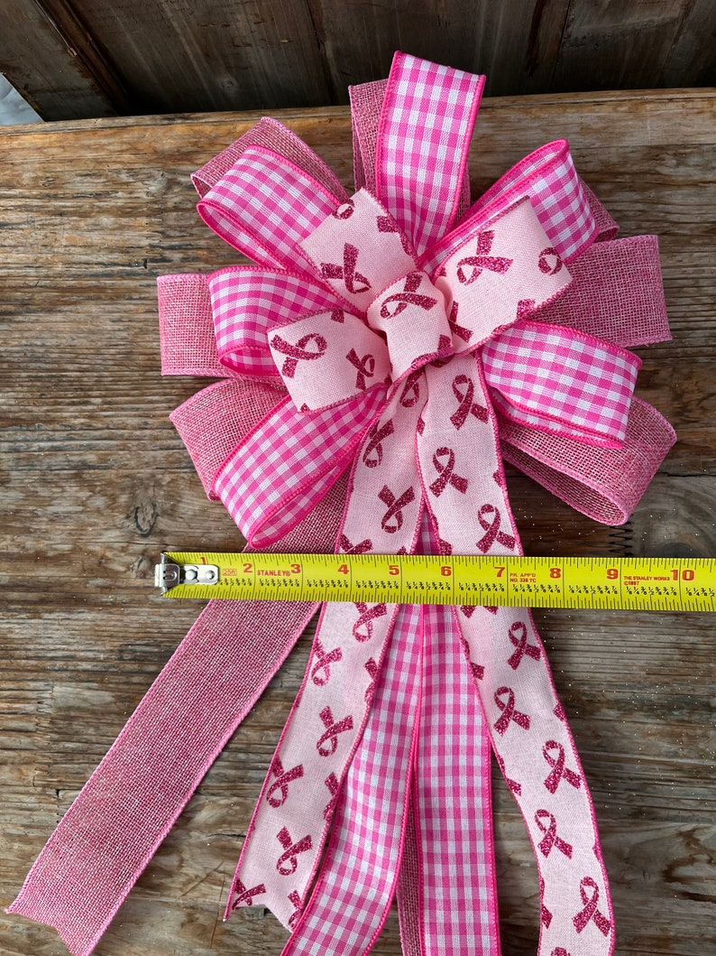 May include: Pink and white gingham ribbon bow with pink ribbon accents featuring a pink ribbon design. The bow is tied with a pink burlap ribbon and measures approximately 10 inches in length.