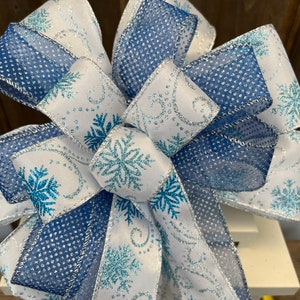 May include: A decorative bow featuring white and blue ribbons with snowflake and swirl patterns. The bow has a layered design with a mix of solid and patterned ribbons, perfect for winter or holiday decor.