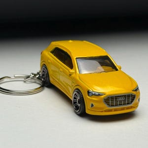 Puede incluir: Un llavero de coche modelo amarillo. El SUV en miniatura tiene ruedas negras y un anillo de llavero plateado. El coche presenta una parrilla delantera y faros detallados. El fondo es un degradado de blanco a negro.