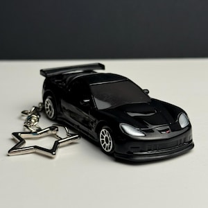 Könnte beinhalten: Ein schwarzes Spielzeugauto mit Spoiler und silbernen Felgen, befestigt an einem silbernen sternförmigen Schlüsselanhänger. Das Auto ist eine Miniaturreplik eines Sportwagens, vor weißem Untergrund und schwarzem Hintergrund.