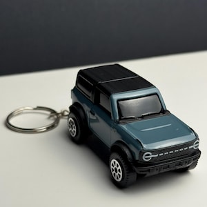Może przedstawiać: Brelok do kluczy z zabawką SUV w kolorze morskim i czarnym. Miniaturowy pojazd ma czarne koła i czarny dach. Kółko breloka jest srebrne. Z przodu pojazdu znajduje się napis "BRONCO" w kolorze białym.