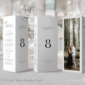 May include: A white tri-fold table number card with a black number 8, the text "Table" and "The Menu" in black script font, and the text "MARCY JACOB 05.25.2025". The card has a photo of a couple in a forest on the back.