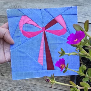 May include: A square, handmade quilt block featuring a geometric design of a gift. The quilt block has a light blue background with a pink bow and dark red ribbon. The block is held by a hand, with purple flowers in the background.