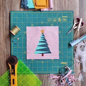 Könnte beinhalten: Ein Weihnachtsbaum-Quiltblock in Rosa, Türkis und Gold, mit einem Stern oben, liegt auf einer grünen Schneidematte. Nähwerkzeuge wie Schere, Rollschneider und Stecknadeln umgeben den Block. Stoffreste in verschiedenen Farben sind verstreut.