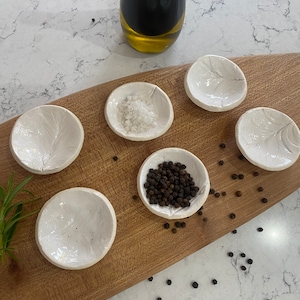 May include: Six white ceramic bowls with a leaf design on a wooden board. One bowl contains salt, another contains black peppercorns, and the other four are empty. A bottle of olive oil is in the background.