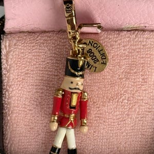 May include: Gold-tone charm featuring a red and white nutcracker figurine with a black hat and gold accents. The charm has a gold tag that reads "Limited Edition 2006".