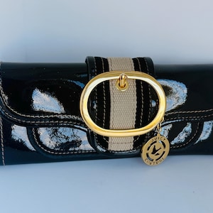 May include: A black patent leather clutch bag with a gold-tone buckle and a beige stripe. The bag has a gold charm with the words "Juicy Couture" and a logo. The edges are stitched.