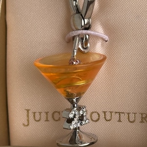 Peut inclure: Un charme argenté avec un verre à martini en verre orange transparent avec un agitateur en argent et un strass rose. Le verre a une base argentée avec le numéro 21 décoré de strass. Le charme est attaché à un fermoir à homard argenté. Le charme est sur un fond rose avec le texte "Juicy Couture" visible.