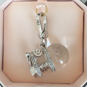 May include: A silver charm with a lobster clasp and a small, round, clear charm with the word "Cherie" etched on it. The charm has a rectangular shape with a crown design and is encrusted with small, clear stones.