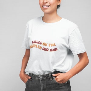 Puede incluir: Camiseta blanca con un gráfico de estilo retro que dice "Eagles on the Warpath 000 ahh" en marrón y naranja.