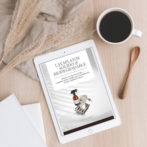 May include: A digital tablet displaying an ebook cover with the title "Lavaplatos Sólido & Biodegradable" in black text. The cover features a white background with a light grey overlay and a photo of a clear glass jar, a white bar of soap, and a spray bottle with a green cap.