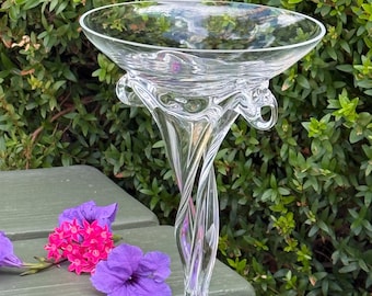 May include: A clear glass goblet featuring a wide, flared bowl and a decorative, twisted stem design. The glass has a smooth, transparent finish with intricate swirling patterns integrated into the structure of the stem and base.