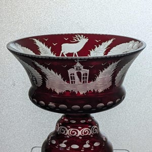 May include: A red glass bowl with a white deer and a white building design. The bowl has a white rim and a white base with a swirl design.
