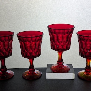 May include: Four ruby red stemmed glasses with a diamond pattern. The glasses have a flared bowl and a yellow-tinted stem and base. The glasses are arranged in a row against a neutral background.