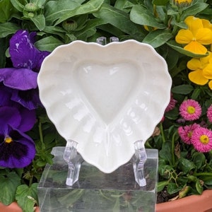 May include: White porcelain heart-shaped dish with a fluted edge. The dish is on a clear acrylic stand.