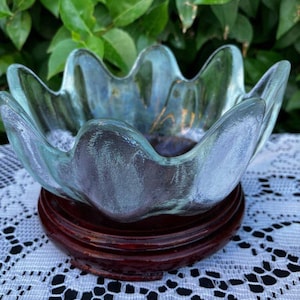 May include: A decorative glass bowl with a scalloped rim, resting on a dark wooden stand. The bowl has a blue-green hue and sits on a round, dark brown wooden base. The background features green foliage.