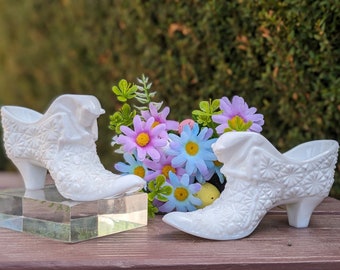 May include: Two white milk glass slipper-shaped vases with a floral design. The vases are sitting on a clear glass surface with a bouquet of purple and blue flowers behind them.