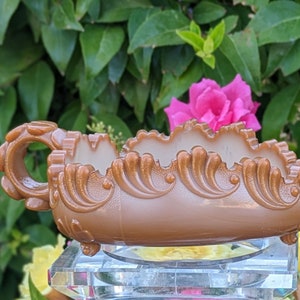 May include: A brown, ornate, antique-style dish with a handle. The dish has a scalloped edge and sits on a clear, faceted base. The background features green foliage and a pink rose.