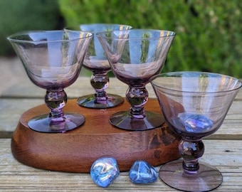 May include: Four vintage amethyst-colored cocktail glasses with a round base and a bulbous stem, displayed on a wooden serving tray. Two blue decorative stones are in front of the tray. The glasses have a flared bowl shape.