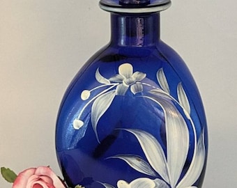 May include: A cobalt blue glass decanter with a matching stopper. The decanter is decorated with white floral designs. A pink and yellow rose is next to the decanter.