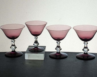 May include: Four vintage stemmed cocktail glasses. Each glass features a wide, shallow bowl in a dusty rose hue, supported by a clear, faceted stem and a dark purple base. The glasses are arranged in a row, showcasing their elegant design.