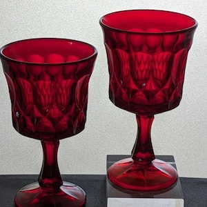 May include: Two ruby red goblets with a detailed, textured pattern. The stemware features a wide bowl and a pedestal base. The glasses are displayed on a black surface with a clear acrylic block. The goblets are made of glass.