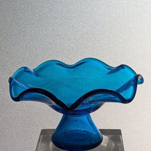 May include: A blue glass bowl with a ruffled edge sits on a pedestal. The bowl is made of textured glass and has a unique, organic shape.