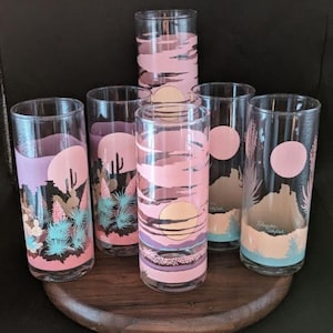 May include: Set of six vintage highball glasses with a desert landscape design. Each glass features a pink and purple sunset scene with cacti and a large sun. The glasses are arranged on a round wooden tray.