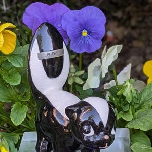 May include: Black and white ceramic skunk figurine with a shiny glaze. The skunk has a raised tail with the word "PHEW" written on it. The figurine is surrounded by purple and yellow flowers.