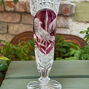 May include: A clear crystal vase with a ruby red overlay, featuring an etched bird and leaf design. The vase has a flared top with a cut crystal pattern and a round base. The vase is approximately 10 inches tall.