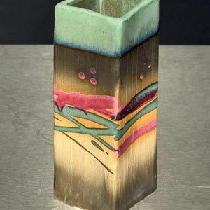 May include: A square ceramic vase featuring a light green rim that transitions into a dark brown body. The surface is decorated with horizontal bands of pink, teal, and yellow, accented by small pink dots and abstract teal line patterns.