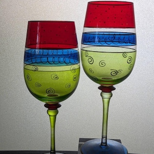 May include: Two hand-painted wine glasses. The rims are red with black dots, followed by blue stripes and green bodies with black swirls. The stems are green with red accents, and the bases are blue.