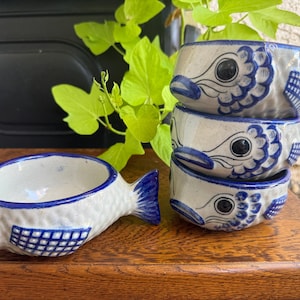 May include: A set of four ceramic bowls shaped like fish, featuring a cream-colored base with hand-painted blue details including scales, fins, and eyes. One bowl is positioned horizontally, while the other three are stacked vertically.
