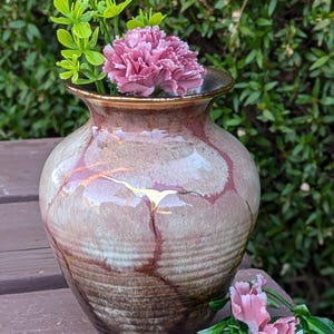 May include: A ceramic vase with a crackled glaze in shades of brown, pink, and gold. The vase is filled with pink carnations and green foliage.