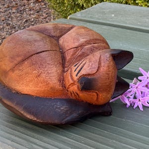 May include: A hand-carved wooden figurine of a sleeping cat with a smooth, polished finish. The sculpture features natural brown wood grain tones, with darker stained areas on the ears and tail, accompanied by a small cluster of light purple flowers.