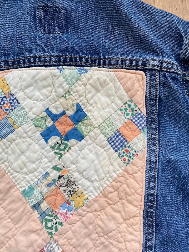 May include: Close-up of a denim jacket with a quilted patch. The patch features a patchwork design with squares of various patterns and colors, including blue, green, orange, and floral prints. The quilt is set against a light pink background.