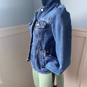 May include: A classic blue denim jacket with a button-up front, chest pockets, and a collar. The jacket is displayed on a light green dress form against a light blue wall. The jacket has a vintage look.