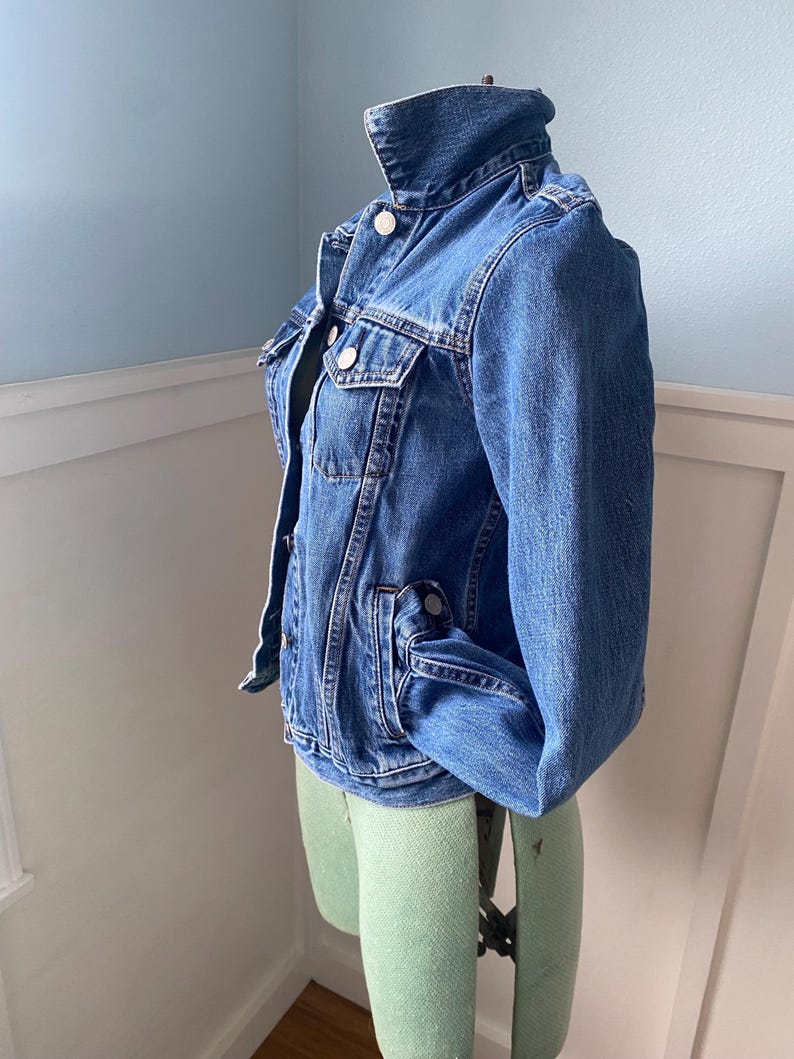 May include: A classic blue denim jacket with a button-up front, chest pockets, and a collar. The jacket is displayed on a light green dress form against a light blue wall. The jacket has a vintage look.