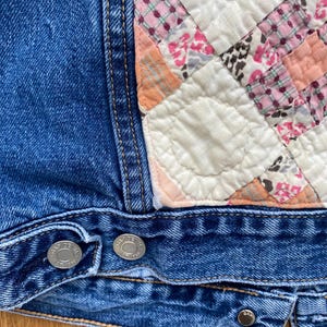 May include: Close-up of a blue denim jacket with a patchwork quilt detail. The quilt features squares of pink, white, and peach fabrics. Silver buttons secure the jacket's front.