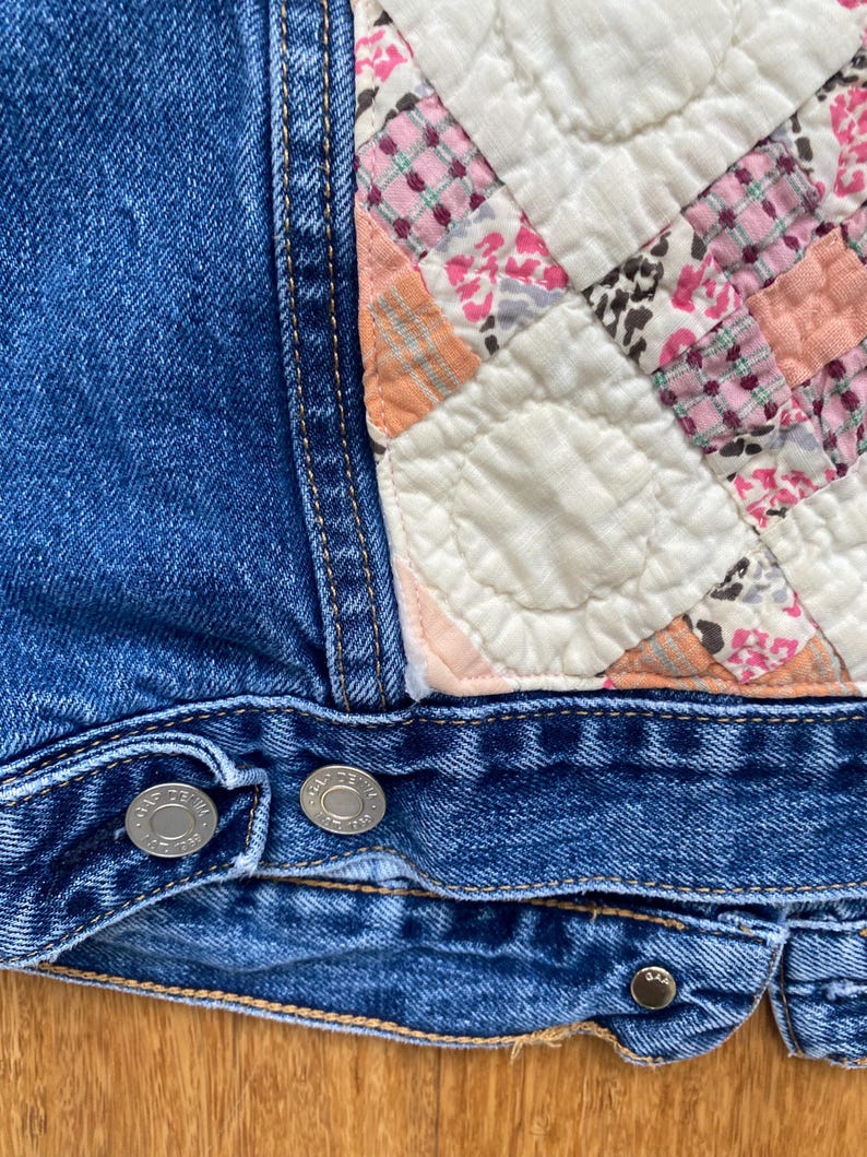 May include: Close-up of a blue denim jacket with a patchwork quilt detail. The quilt features squares of pink, white, and peach fabrics. Silver buttons secure the jacket's front.