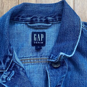 May include: Close-up of a blue denim jacket with the GAP Denim label. The jacket has a button closure and a collar. The size is XS. The denim jacket is a classic wardrobe staple.