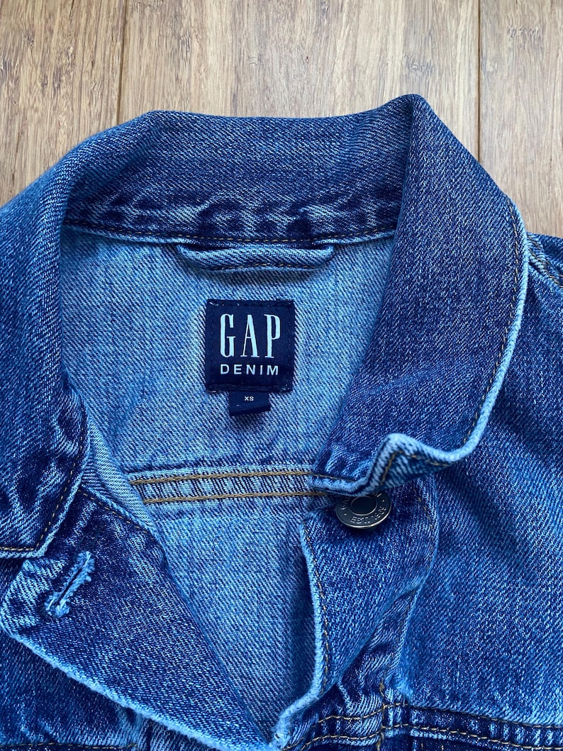 May include: Close-up of a blue denim jacket with the GAP Denim label. The jacket has a button closure and a collar. The size is XS. The denim jacket is a classic wardrobe staple.