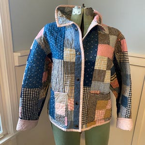 May include: A patchwork jacket with a variety of fabric patterns in blue, pink, and tan. The jacket has a quilted design, a collar, and button closures. The cuffs and trim are a soft pink color. The jacket is displayed on a dress form.
