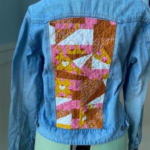 May include: Light blue denim jacket with a colorful patchwork design on the back. The patchwork features geometric shapes in shades of pink, yellow, brown, and white, creating a unique and artistic look. The jacket has a classic collar and button closures.