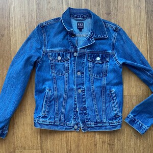 May include: A blue denim jacket with a button-up front, two chest pockets, and two side pockets. The jacket has a classic collar and long sleeves. The brand name "GAP" is visible on the collar. The jacket is made of a medium-wash denim.