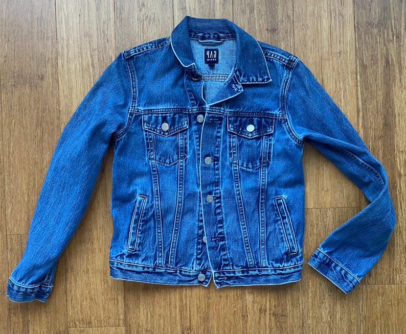 May include: A blue denim jacket with a button-up front, two chest pockets, and two side pockets. The jacket has a classic collar and long sleeves. The brand name "GAP" is visible on the collar. The jacket is made of a medium-wash denim.