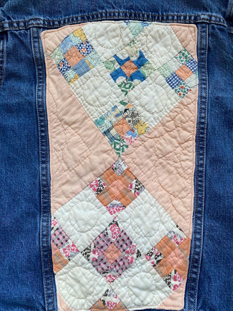 May include: A denim jacket with a quilted back panel. The panel features a patchwork design with squares of various patterned fabrics in shades of blue, pink, orange, and white. The jacket is a medium wash denim.